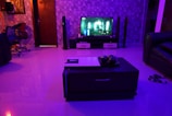 A modern living room is dimly lit with blue and purple ambient lighting. A flat-screen TV displays a scene from a movie or video game. Surround sound speakers are placed around the room. The floor is glossy, reflecting the lights. A dark coffee table in the center of the room holds decorative objects. To the left, a comfortable chair sits near a wall with a geometric pattern. There is a pair of sandals on the floor near a sofa.