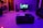 A modern living room is dimly lit with blue and purple ambient lighting. A flat-screen TV displays a scene from a movie or video game. Surround sound speakers are placed around the room. The floor is glossy, reflecting the lights. A dark coffee table in the center of the room holds decorative objects. To the left, a comfortable chair sits near a wall with a geometric pattern. There is a pair of sandals on the floor near a sofa.