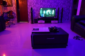 A modern living room is dimly lit with blue and purple ambient lighting. A flat-screen TV displays a scene from a movie or video game. Surround sound speakers are placed around the room. The floor is glossy, reflecting the lights. A dark coffee table in the center of the room holds decorative objects. To the left, a comfortable chair sits near a wall with a geometric pattern. There is a pair of sandals on the floor near a sofa.