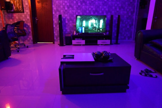 A modern living room is dimly lit with blue and purple ambient lighting. A flat-screen TV displays a scene from a movie or video game. Surround sound speakers are placed around the room. The floor is glossy, reflecting the lights. A dark coffee table in the center of the room holds decorative objects. To the left, a comfortable chair sits near a wall with a geometric pattern. There is a pair of sandals on the floor near a sofa.