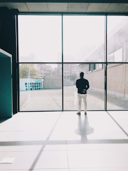 A young professional standing by a window, gazing outside with a hopeful expression in a minimalist office space.
