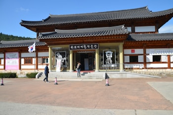 A traditional Korean building with wooden accents and gray tiled roofs stands under a clear blue sky. There are several signs and banners with Korean characters, and the South Korean flag is displayed on a flagpole. Two people are walking near the entrance, which features artistic drawings of masked figures.