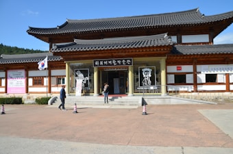 A traditional Korean building with wooden accents and gray tiled roofs stands under a clear blue sky. There are several signs and banners with Korean characters, and the South Korean flag is displayed on a flagpole. Two people are walking near the entrance, which features artistic drawings of masked figures.