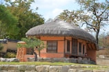 A traditional house with a thatched roof and orange walls surrounded by trees and stone walls. The structure has wooden beams and large windows. A small tree is situated at its front near a stone path.