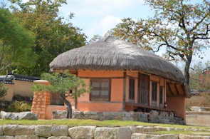 A traditional house with a thatched roof and orange walls surrounded by trees and stone walls. The structure has wooden beams and large windows. A small tree is situated at its front near a stone path.