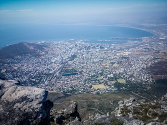Aerial view of a cityscape featuring a dense urban area with numerous buildings, sprawling residential neighborhoods, and a large bay. The coastline curves around the city, creating a natural harbor. Foreground includes rocky terrain and sparse vegetation.