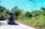 A jeepney is traveling on a rural road surrounded by lush greenery and tall trees. Several people are sitting on top of the vehicle enjoying an open-air ride. A single street lamp is visible on the side of the road, and the sky is clear and blue.
