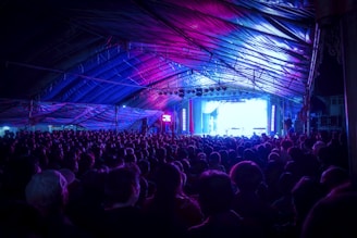 A large audience is gathered inside a tent-like structure, all facing towards a brightly lit stage area at the front. The atmosphere is vibrant with a striking combination of purple, blue, and pink lighting that fills the venue. The ceiling and sides are covered with reflective materials that catch these colorful lights, adding to the ambiance.