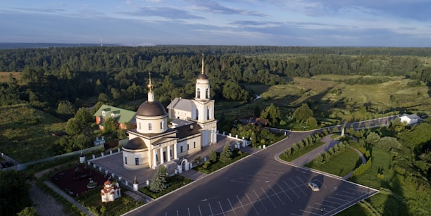 A large church building with domes and a bell tower surrounded by a lush green landscape. The architecture is accented with gold and white colors. An expansive parking lot in the foreground contains very few cars, suggesting a quiet atmosphere.