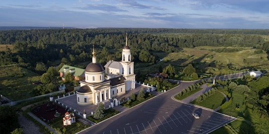 A large church building with domes and a bell tower surrounded by a lush green landscape. The architecture is accented with gold and white colors. An expansive parking lot in the foreground contains very few cars, suggesting a quiet atmosphere.