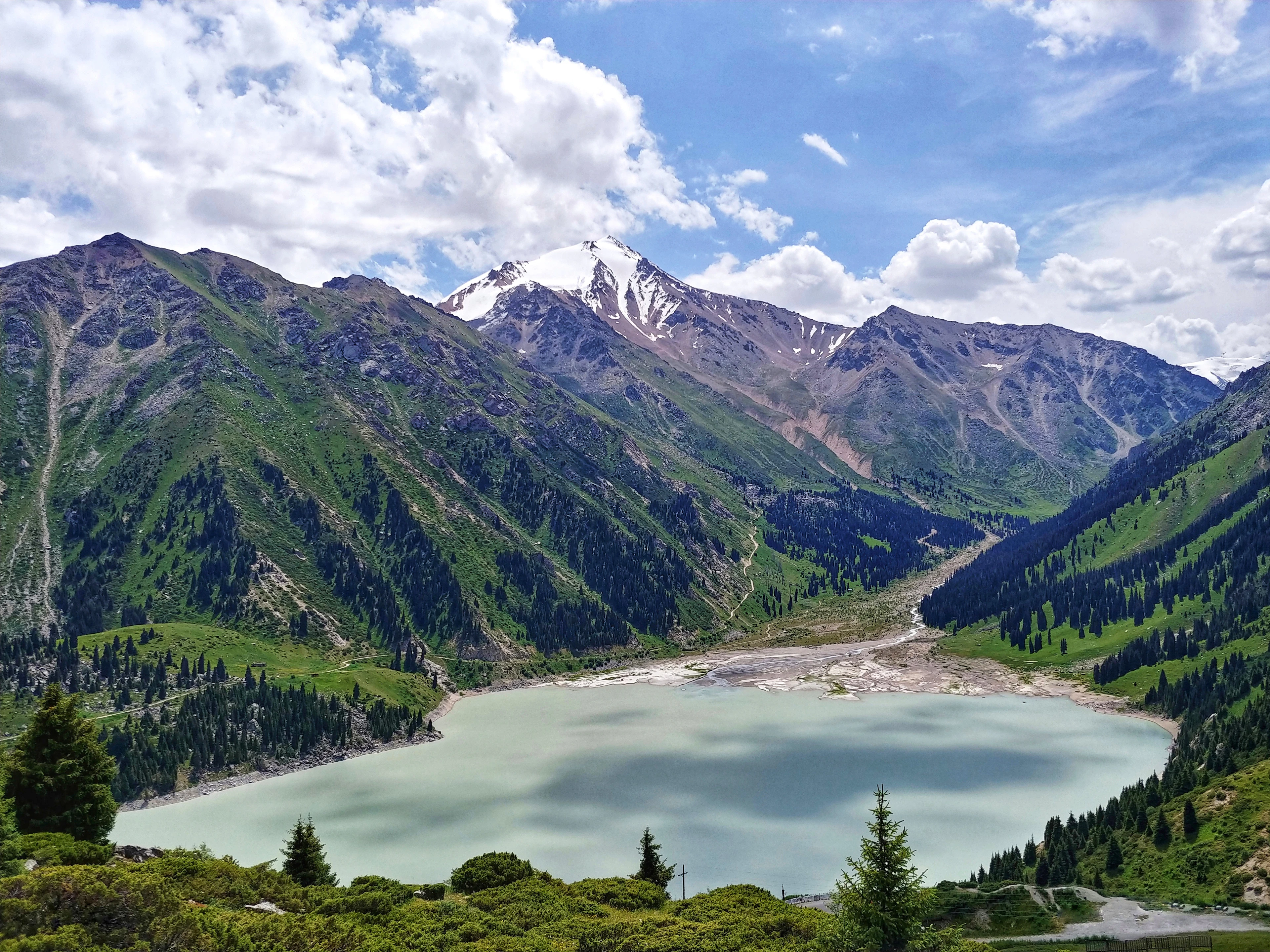 A turquoise mountain lake sits beneath a snow-capped peak under a partly cloudy sky.