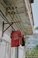 A clothesline is mounted on a weathered concrete wall under a deteriorating roof. Several shirts in red, brown, and black hang from hangers. Electric wiring is visible on the wall, and shadows of the shirts are cast on the surface. An adjacent building and some greenery are seen in the background.