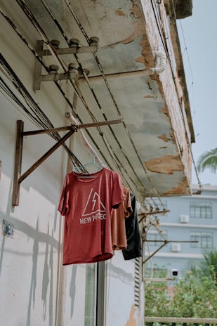 A clothesline is mounted on a weathered concrete wall under a deteriorating roof. Several shirts in red, brown, and black hang from hangers. Electric wiring is visible on the wall, and shadows of the shirts are cast on the surface. An adjacent building and some greenery are seen in the background.