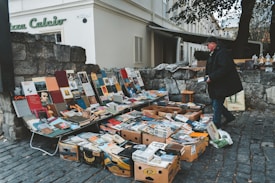 A street vendor is selling a collection of books and magazines displayed on tables and in boxes. The setup is on a cobblestone street next to a stone wall, with various colorful book covers visible. The vendor, dressed in a dark coat and cap, is interacting with the displayed items. In the background, there is an old building with shutters and a sign that reads 'Piazza Calcio'.