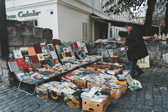 A street vendor is selling a collection of books and magazines displayed on tables and in boxes. The setup is on a cobblestone street next to a stone wall, with various colorful book covers visible. The vendor, dressed in a dark coat and cap, is interacting with the displayed items. In the background, there is an old building with shutters and a sign that reads 'Piazza Calcio'.