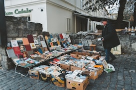 A street vendor is selling a collection of books and magazines displayed on tables and in boxes. The setup is on a cobblestone street next to a stone wall, with various colorful book covers visible. The vendor, dressed in a dark coat and cap, is interacting with the displayed items. In the background, there is an old building with shutters and a sign that reads 'Piazza Calcio'.