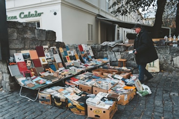 A street vendor is selling a collection of books and magazines displayed on tables and in boxes. The setup is on a cobblestone street next to a stone wall, with various colorful book covers visible. The vendor, dressed in a dark coat and cap, is interacting with the displayed items. In the background, there is an old building with shutters and a sign that reads 'Piazza Calcio'.