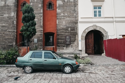 A green sedan car is parked on a cobblestone surface next to a building with a mix of brick and stucco walls. The building features red accents around the windows and a large wooden door leads into an arched entryway. A tall, narrow evergreen tree is positioned beside the car. A plaque and some small plants are visible on the building's exterior.