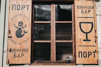 Wooden window shutters with signs and illustrations painted in black. The left shutter features a mermaid holding a glass, with text in a foreign language, while the right shutter displays a large wine glass icon and more text.
