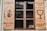 Wooden window shutters with signs and illustrations painted in black. The left shutter features a mermaid holding a glass, with text in a foreign language, while the right shutter displays a large wine glass icon and more text.