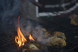 Grilled pieces of meat are sizzling on a barbecue with flames and smoke rising from the heat. The meat appears to be marinated or seasoned, with visible char and grill marks on some parts.