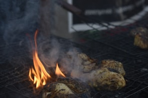 Grilled pieces of meat are sizzling on a barbecue with flames and smoke rising from the heat. The meat appears to be marinated or seasoned, with visible char and grill marks on some parts.
