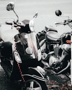 A close-up view of a black scooter and a motorcycle parked side by side. The scooter has a reflective surface, chrome mirrors, and a vaguely visible red cable coiled around it. The motorcycle has a visible engine and chrome elements. Fallen leaves are scattered on the ground beneath them.