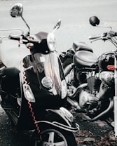 A close-up view of a black scooter and a motorcycle parked side by side. The scooter has a reflective surface, chrome mirrors, and a vaguely visible red cable coiled around it. The motorcycle has a visible engine and chrome elements. Fallen leaves are scattered on the ground beneath them.