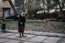 An elderly woman is sweeping a paved sidewalk covered with scattered leaves. She is wearing a dark sweater, red skirt, and brown boots. Behind her, there are trees with sparse foliage and a playground area with swings and people sitting. A weathered building and a black car are visible in the background.