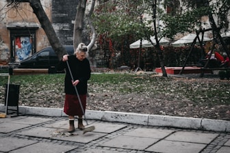 An elderly woman is sweeping a paved sidewalk covered with scattered leaves. She is wearing a dark sweater, red skirt, and brown boots. Behind her, there are trees with sparse foliage and a playground area with swings and people sitting. A weathered building and a black car are visible in the background.
