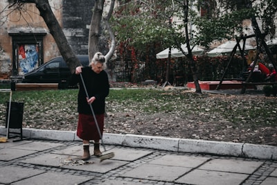 An elderly woman is sweeping a paved sidewalk covered with scattered leaves. She is wearing a dark sweater, red skirt, and brown boots. Behind her, there are trees with sparse foliage and a playground area with swings and people sitting. A weathered building and a black car are visible in the background.