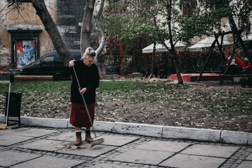 An elderly woman is sweeping a paved sidewalk covered with scattered leaves. She is wearing a dark sweater, red skirt, and brown boots. Behind her, there are trees with sparse foliage and a playground area with swings and people sitting. A weathered building and a black car are visible in the background.