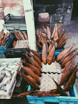 Numerous smoked fish are arranged in blue crates, displayed at an outdoor market, with additional crates containing fresh fish nearby. A weathered stone wall is in the background alongside a chair with a wooden board resting on it.