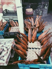 Numerous smoked fish are arranged in blue crates, displayed at an outdoor market, with additional crates containing fresh fish nearby. A weathered stone wall is in the background alongside a chair with a wooden board resting on it.