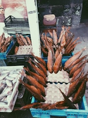 Numerous smoked fish are arranged in blue crates, displayed at an outdoor market, with additional crates containing fresh fish nearby. A weathered stone wall is in the background alongside a chair with a wooden board resting on it.