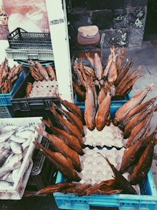 Numerous smoked fish are arranged in blue crates, displayed at an outdoor market, with additional crates containing fresh fish nearby. A weathered stone wall is in the background alongside a chair with a wooden board resting on it.