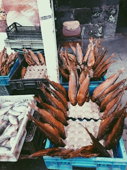 Numerous smoked fish are arranged in blue crates, displayed at an outdoor market, with additional crates containing fresh fish nearby. A weathered stone wall is in the background alongside a chair with a wooden board resting on it.