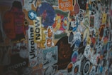 A colorful collage of various stickers on a surface, including images of cartoon characters, animals, and text-based designs in multiple languages. Each sticker has a unique design that ranges from simple shapes to detailed illustrations with humorous and artistic themes.