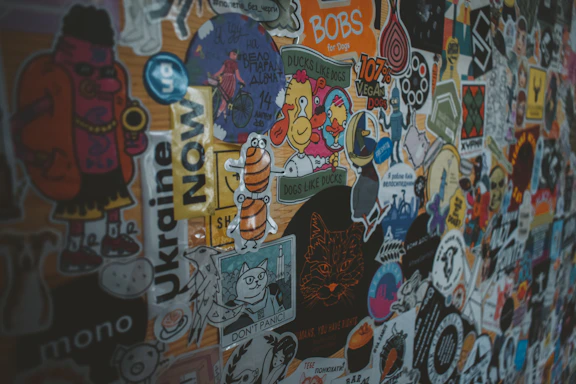 A colorful collage of various stickers on a surface, including images of cartoon characters, animals, and text-based designs in multiple languages. Each sticker has a unique design that ranges from simple shapes to detailed illustrations with humorous and artistic themes.