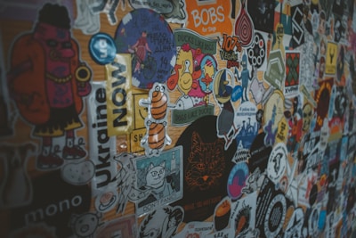 A colorful collage of various stickers on a surface, including images of cartoon characters, animals, and text-based designs in multiple languages. Each sticker has a unique design that ranges from simple shapes to detailed illustrations with humorous and artistic themes.