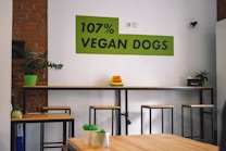 A modern interior featuring a wooden bar table with several metal stools. A green plant is placed on a table's end, and a bright green sign on the wall reads '107% VEGAN DOGS'. The setting includes exposed brick walls and minimalistic decor, creating a contemporary atmosphere.