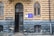 A vintage architectural facade with ornate windows and a central door featuring intricate metalwork designs. The building’s exterior is composed of textured beige and dark gray brickwork, and there are visible signs of wear. A blue and gold sign is mounted between the windows.