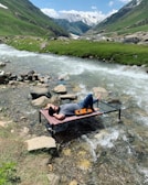 A person relaxes on a small bed placed over a stream in a mountainous landscape. The clear water flows beneath, surrounded by rocks and lush green grass. In the background, the scene extends to snow-capped mountains under a blue sky with scattered clouds.