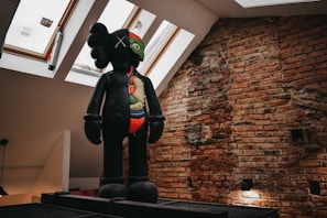 A large, stylized sculpture depicting a figure with a multi-colored face and visible internal organs stands against an exposed brick wall. The figure appears to be positioned on a platform in a room with skylight windows, allowing natural light to illuminate the scene.