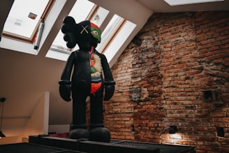 A large, stylized sculpture depicting a figure with a multi-colored face and visible internal organs stands against an exposed brick wall. The figure appears to be positioned on a platform in a room with skylight windows, allowing natural light to illuminate the scene.