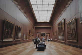 A grand art gallery with high ceilings and wooden parquet floors, displaying numerous large framed paintings on both walls. Several people are casually observing the artwork, while others are seated on a bench in the center of the room.