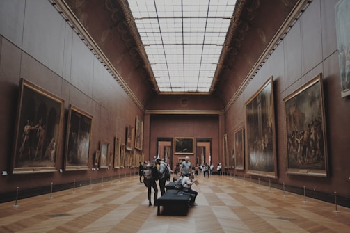 A grand art gallery with high ceilings and wooden parquet floors, displaying numerous large framed paintings on both walls. Several people are casually observing the artwork, while others are seated on a bench in the center of the room.
