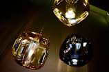 Three modern pendant lights with glass shades in varying tones of amber and black. The lighting fixtures are reflected on a shiny surface, possibly metallic, creating an abstract visual effect.