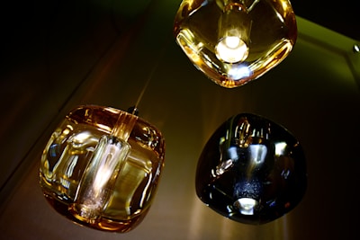 Three modern pendant lights with glass shades in varying tones of amber and black. The lighting fixtures are reflected on a shiny surface, possibly metallic, creating an abstract visual effect.