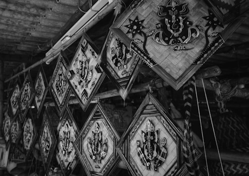 A collection of intricately designed, diamond-shaped artworks hanging in a row. Each piece features a central pattern resembling an abstract or cultural symbol, set against a woven backdrop. The scene conveys a rustic, artisanal atmosphere with wooden beams and other handmade items visible.
